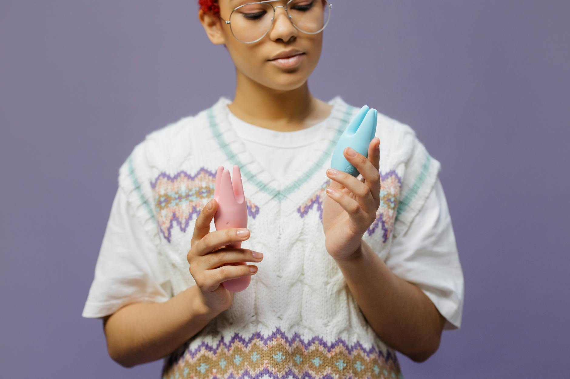A woman with eyeglasses holding a blue clitoral vibrator in a contemplative, curious manner against a neutral background.