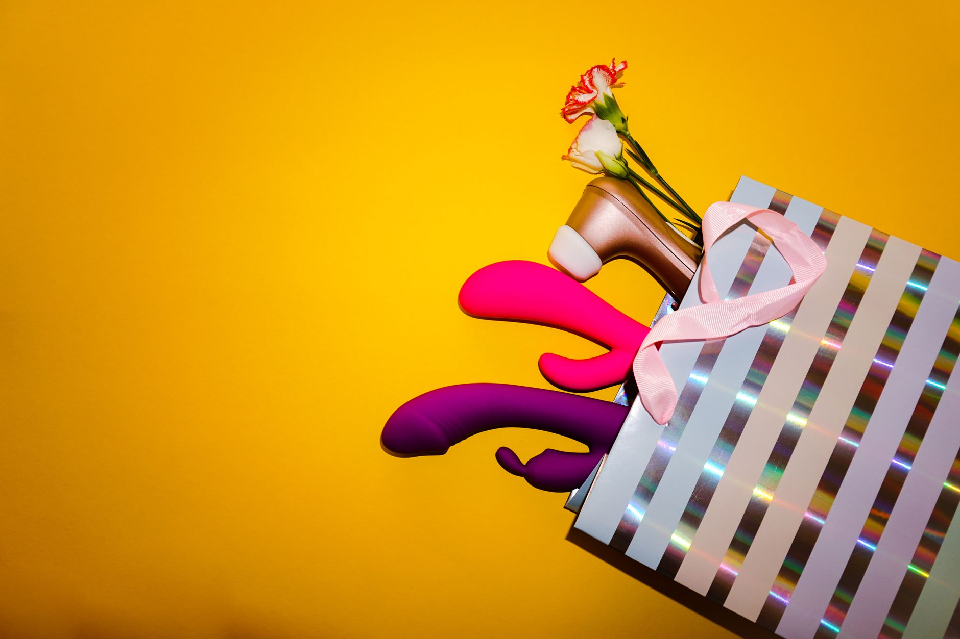Vibradores coloridos com flores em uma bolsa de presente holográfica, em fundo amarelo vibrante.